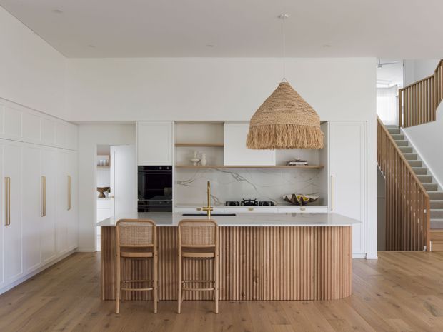 Bright open-plan kitchen with white shaker-style cabinetry, brass hardware, a white stone benchtop and splashback, integrated black appliances, and a fluted timber island with woven bar stools. A large woven pendant light hangs above the island, while a timber staircase with vertical balustrading is visible to the right. Warm timber flooring and neutral finishes create a refined, coastal-inspired interior.