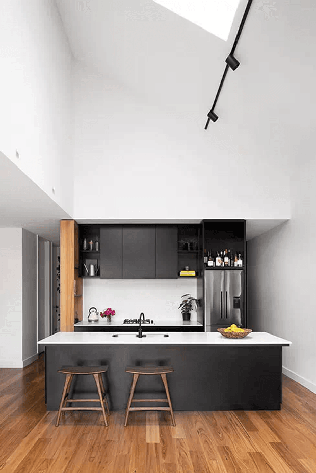 Contemporary open-plan kitchen viewed from the living area, featuring a large dark island with white benchtop and seating for two, matching dark overhead cabinetry, integrated stainless-steel refrigerator, timber flooring, and a high white ceiling with black track lighting.