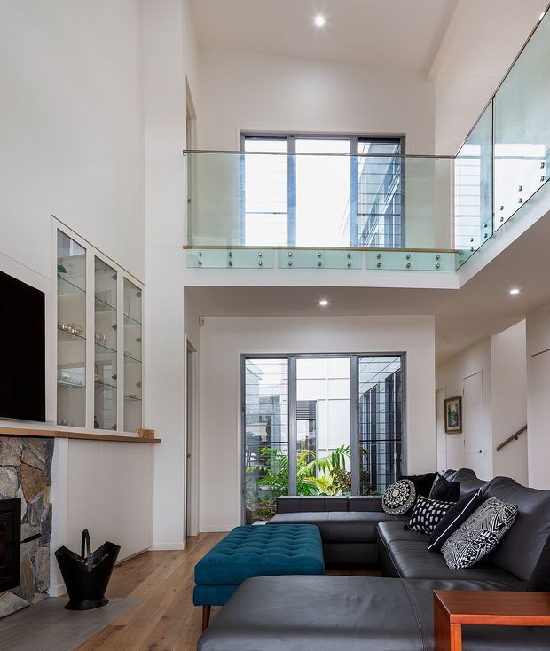 Bright contemporary double-height living room with a mezzanine-level glazed balustrade, large windows, and an internal courtyard view. The room features a black leather sectional sofa, teal upholstered ottoman, timber flooring, a stone-clad fireplace, built-in glass display cabinetry, and recessed ceiling lights.