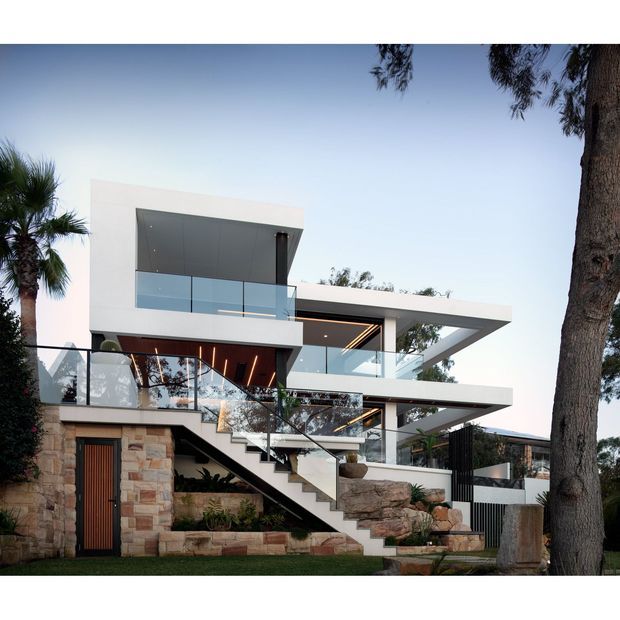 A striking multi-level contemporary residence with a strong geometric composition, white rendered volumes, broad cantilevered roof planes, extensive glass balustrades, and exposed external stairs. The lower level combines stacked stone cladding with a timber entry door, while landscaped rockwork and mature trees frame the property.