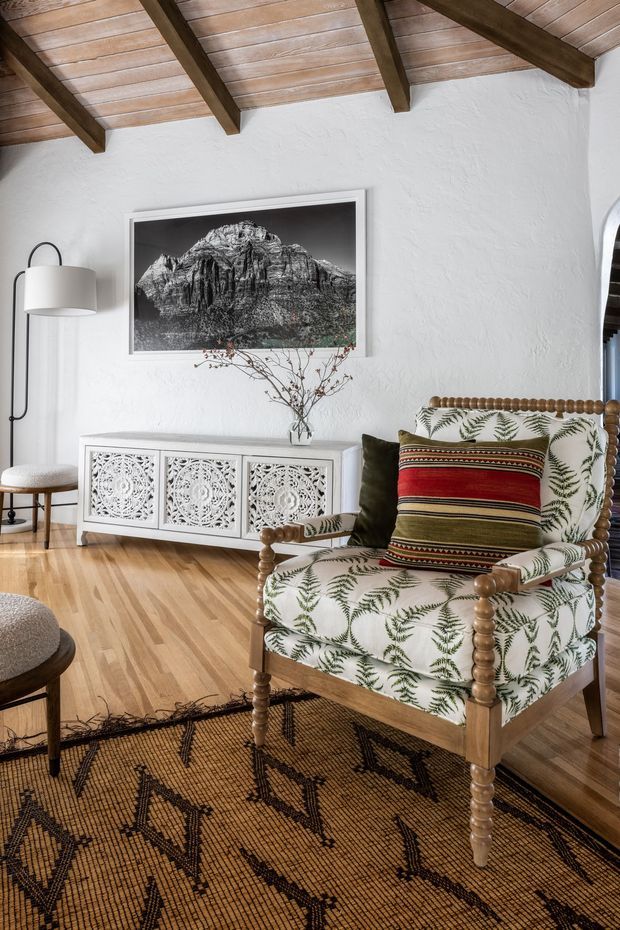 A warmly styled living room with white textured plaster walls, a timber-lined vaulted ceiling with exposed dark beams, and light timber flooring. A patterned upholstered armchair with turned wooden detailing and a striped cushion sits on a large geometric woven rug. A white decorative sideboard, floor lamp, mountain photograph, and small vase complete the room.