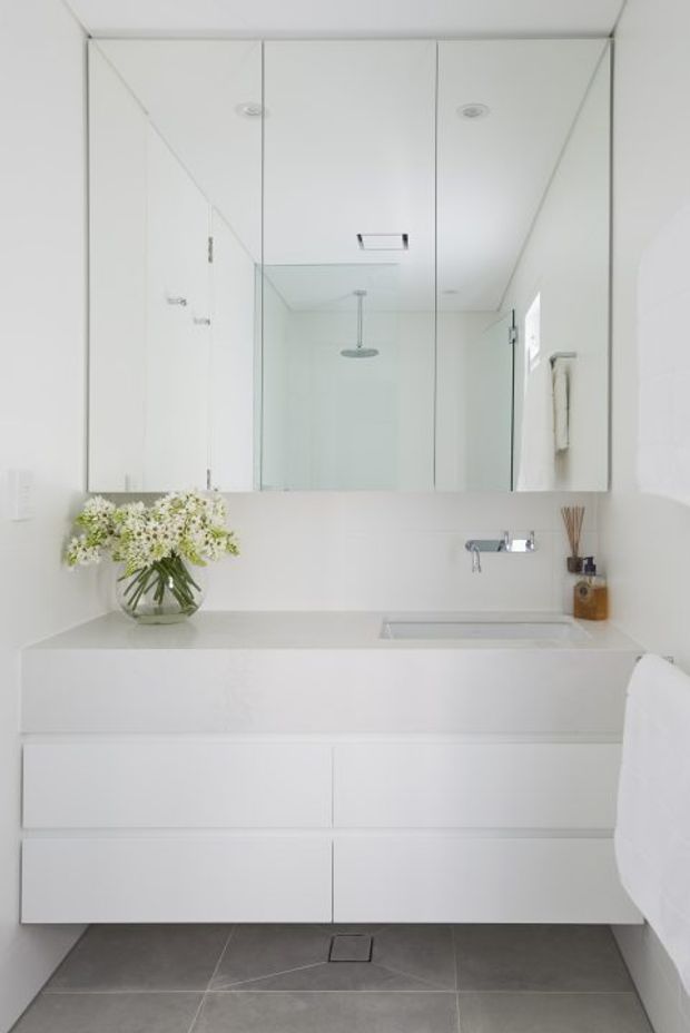 Bright minimalist bathroom featuring a wall-mounted white vanity with integrated basin, large mirrored wall cabinets, a wall-mounted chrome tap, and a frameless glass shower visible in the reflection. Grey tiled flooring provides contrast, while simple styling includes a vase of flowers and a reed diffuser.