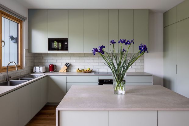 Contemporary kitchen featuring pale grey-green flat-panel cabinetry, a large stone island with waterfall-style edges, a light subway-tile backsplash, dark timber flooring, and timber-framed glazing. A vase of purple flowers sits on the island, with integrated appliances and understated modern fixtures creating a calm, refined interior.