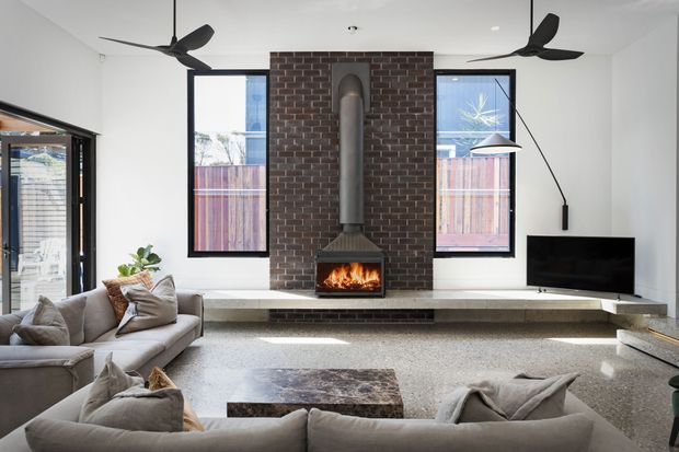 Bright contemporary living room centered on a tall exposed brick fireplace with a suspended metal flue and built-in hearth bench. The room features white walls and ceiling, large black-framed windows and glazing, ceiling fans, a neutral sectional sofa, a low coffee table, and a wall-mounted television.