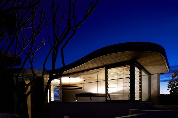 Twilight exterior view of a low, sculptural modern building with a sweeping curved roof and extensive illuminated glazing. Timber-toned soffits and horizontal louvres create a warm contrast against the dark facade and deep blue sky, while silhouetted trees frame the foreground.