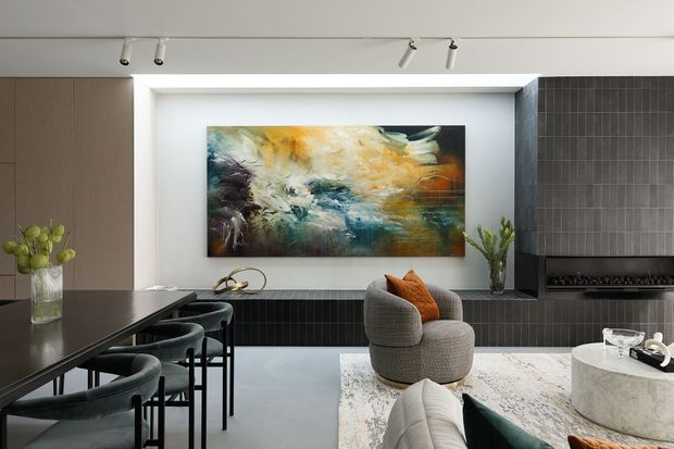 A refined contemporary open-plan living and dining area with a dark kitchen island and bar stools in the foreground, a sculptural grey lounge chair and sofa, and a large circular stone coffee table. The room features a dramatic dark tiled fireplace wall, a recessed display niche with abstract artwork, neutral cabinetry, track lighting, and a pale patterned rug.