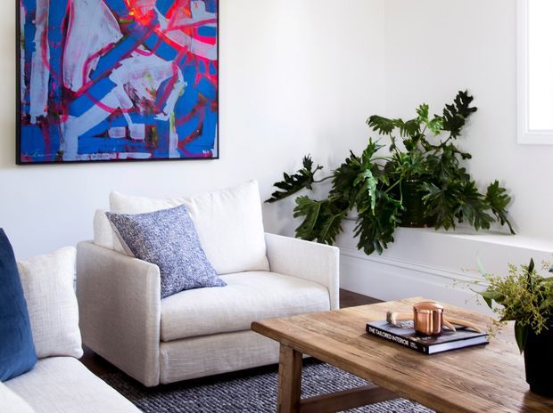 Bright, refined living room featuring a white upholstered armchair and partial sofa with blue and patterned cushions, a rustic timber coffee table, a large vivid abstract artwork, and abundant leafy plants. White wall panelling and a window contribute to the clean, light-filled setting.