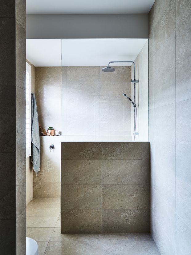A contemporary walk-in bathroom with a frameless glass shower enclosure, wall-mounted rainfall and handheld shower fixtures, and full-height beige-grey stone-look tiling across the walls, floor, and shower partition. A large mirror spans the rear wall, with a small plant and towel adding subtle detail. Natural light enters from a narrow side window, creating a bright, calm atmosphere.