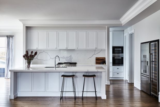 A bright, refined kitchen with pale grey shaker-style cabinetry, a large island with seating for two, white marble-look splashback and benchtop surfaces, integrated stainless-steel appliances, dark timber flooring, and classic decorative cornices. A glass-front wine or drinks cabinet is visible at right, with a glimpse of an adjacent hallway.