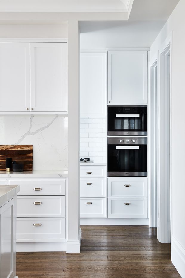 Bright, narrow kitchen view with white shaker-style cabinetry, integrated double wall ovens, white veined stone benchtops and splashback, and dark stained timber flooring. The image looks through a framed opening toward a compact kitchen work zone, with a partial island and timber cutting boards visible at left. Clean, light-filled and highly functional joinery dominates the scene.