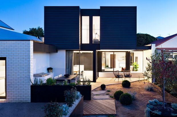 Contemporary two-storey residential home with bold black horizontal cladding, white brickwork, and large glazed openings. The house is arranged around a landscaped courtyard with timber decking, raised garden beds, clipped shrubs, and an outdoor seating area. Warm interior lighting contrasts with the dark facade and blue evening sky.