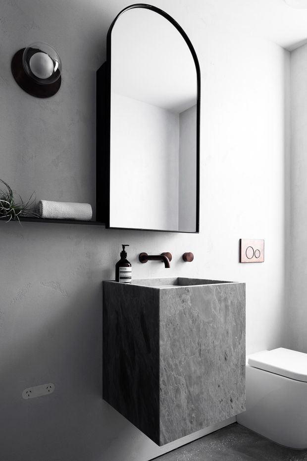 A compact, monochrome contemporary bathroom featuring a sculptural grey stone or concrete vanity basin, wall-mounted dark metal tapware, a large arched black-framed mirror, integrated shelving, wall sconce, and a partially visible white toilet. Textured grey walls and a dark floor create a refined, moody minimalist atmosphere.