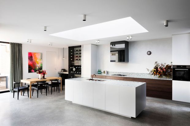 Bright open-plan kitchen and dining area with a large white island, integrated sink, dark timber cabinetry, speckled stone surfaces, built-in oven, wine storage, and a dining table beside full-height curtains. A large rectangular skylight brings natural light into the polished concrete-finished space, with colourful artwork and floral arrangements adding accents.