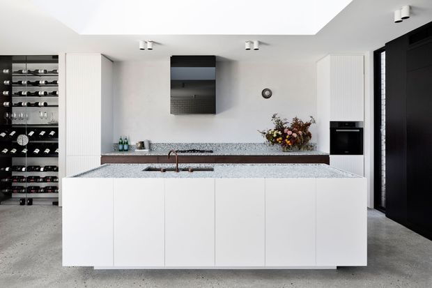 Bright, minimalist contemporary kitchen with a large white island, integrated sink and tap, white flat-panel cabinetry, speckled stone benchtops, a dark tiled splashback, and concealed appliances. A tall wine storage area is visible to the left, with a dark entry door and polished concrete flooring adding contrast.