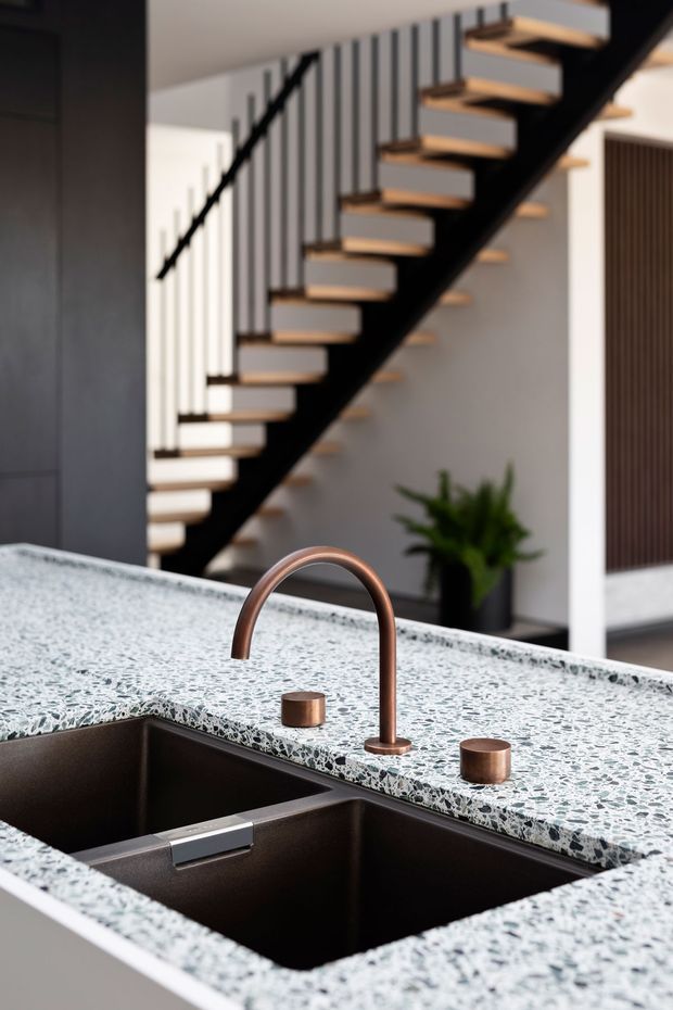 Close-up of a contemporary kitchen island featuring a dark double-bowl undermount sink, curved copper-toned tapware, and matching cylindrical controls set into a terrazzo-style benchtop. A floating timber staircase with black structural elements and vertical balustrade slats is visible in the background.
