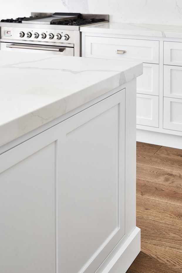 Close-up of a bright, white kitchen featuring a large island with framed shaker-style cabinetry and a white veined stone benchtop. Matching wall cabinetry, a stainless-steel range cooker, and warm timber flooring are visible.