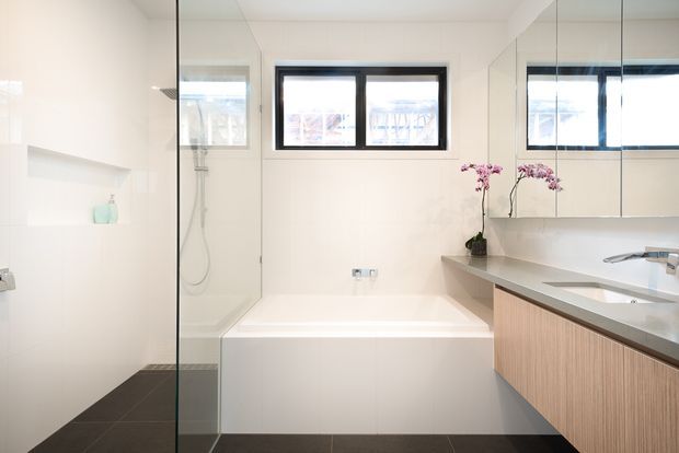 Bright contemporary bathroom with a freestanding-style rectangular bathtub beneath a black-framed window, a separate glass-enclosed shower at left, and a floating timber-look vanity with a pale stone benchtop at right. Mirrored cabinetry and orchid plants add visual depth and a refined finish.