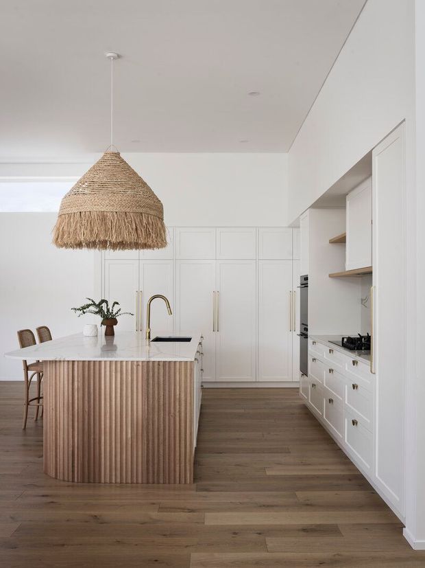 A bright, refined kitchen with white shaker-style cabinetry, integrated appliances, open timber shelving, a pale stone island benchtop, and a fluted timber island front. Warm timber flooring and woven pendant lighting add natural texture, while a brass-toned tap and dining chairs complete the space.