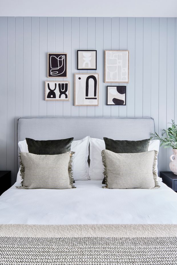 A styled bedroom featuring a large upholstered light-grey bedhead, crisp white bedding, dark charcoal cushions, and striped decorative pillows. The wall behind the bed is clad in pale grey vertical timber panelling and decorated with a curated arrangement of monochrome abstract artwork. Dark bedside tables and a leafy plant complete the calm, contemporary composition.