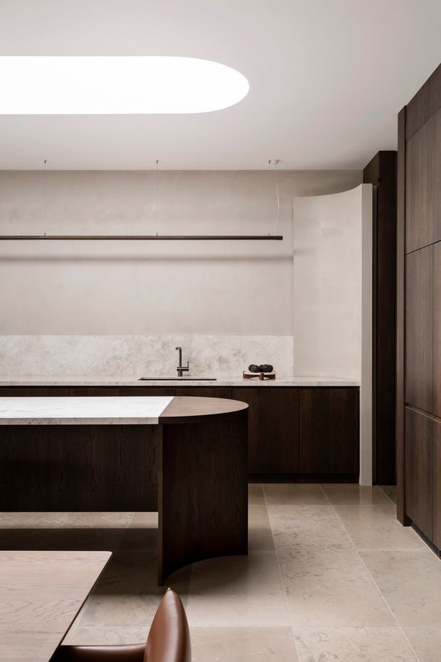 A refined, minimalist kitchen with dark timber cabinetry, a pale stone island and benchtop, integrated tall storage, and a curved island end. Large-format light stone flooring and matching wall surfaces create a warm neutral palette. A linear suspended light and oversized rounded ceiling light add sculptural elements, while a dining table and leather chair are partially visible in the foreground.