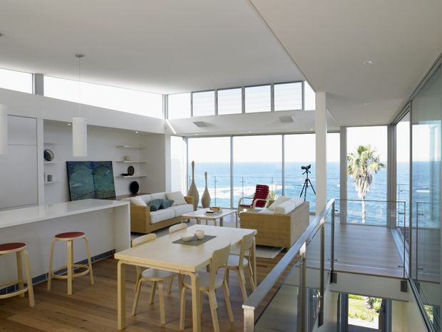 Bright, open-plan coastal home interior with a kitchen, dining area and living room arranged around expansive floor-to-ceiling glazing. The space has white walls and cabinetry, warm timber flooring, light timber dining furniture, woven lounge seating, and a double-height glazed void with a stair balustrade. Ocean views and palms are visible beyond the glazing.