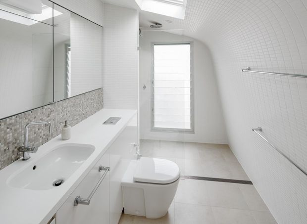 Bright, highly streamlined contemporary bathroom with a wall-hung toilet, white vanity and integrated basin, large mirrored cabinet, mosaic tile splashback, full-height white mosaic wall tiling, and accessible-style stainless steel grab rails. A frosted window and skylight bring in abundant natural light.