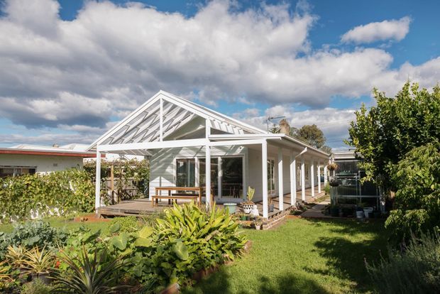 Exterior view of a single-storey white weatherboard house with a prominent gabled porch and open verandah supported by slender white posts. The home has large glazed openings, a low-pitched roof, and a landscaped garden with lawn and established planting in the foreground. Bright daylight and dramatic cloud cover create a welcoming residential setting.
