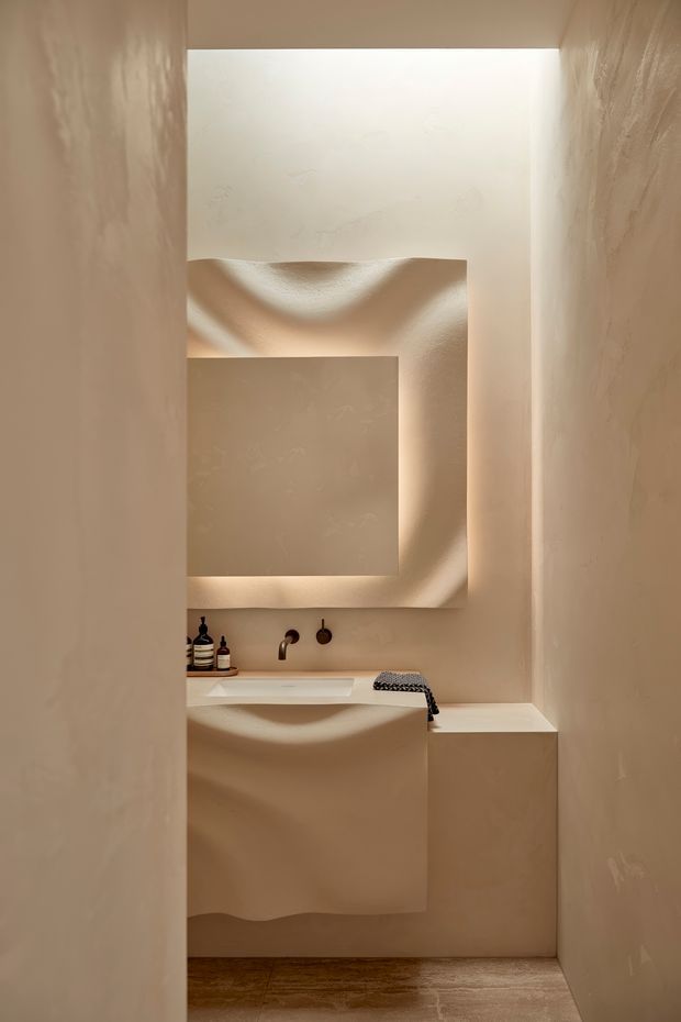 A narrow, sculptural bathroom viewed through a doorway or passage. The room features warm beige textured plaster walls, a minimalist integrated vanity with a white basin, wall-mounted dark bronze or black tapware, a large softly illuminated textured mirror or feature panel, and a built-in bench or ledge. Warm concealed lighting creates curved shadows and an atmospheric, monolithic appearance. The floor appears to be warm timber or wood-look flooring.