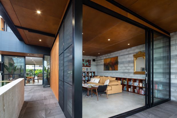 A contemporary open-plan interior centred on a living room viewed through large black-framed glazed sliding doors. The space features exposed grey concrete block walls, extensive warm timber ceiling lining, dark metal framing, built-in low bookcases, a tan sofa, a small lounge chair, artwork and a large ornate gold-framed mirror. A tiled circulation area and planted courtyard are visible beyond the living room.
