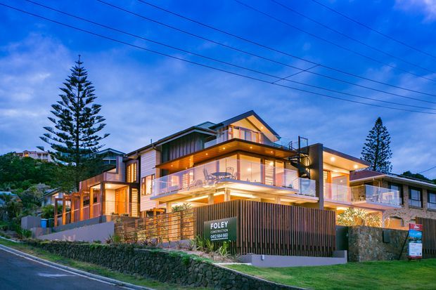 A large contemporary multi-level residential house photographed at dusk from the street. The home features warm illuminated interiors, extensive balconies and glass balustrades, timber and rendered exterior finishes, broad roof overhangs, and landscaped terraced grounds with stone retaining walls. Tall conifer trees and overhead utility lines frame the suburban streetscape.