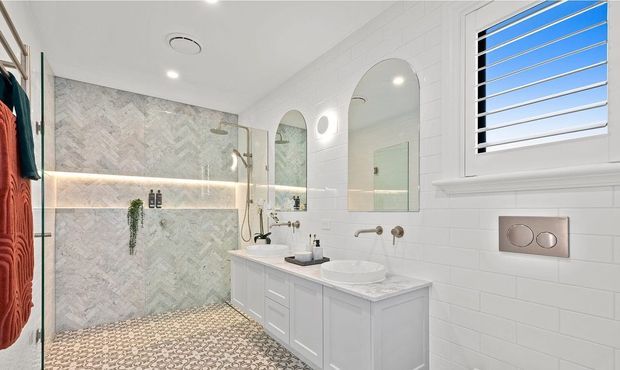 Bright contemporary bathroom featuring a walk-in glass shower with pale herringbone wall tile, a small pebble-pattern floor, a double vanity with white cabinetry and stone benchtop, arched mirrors, wall sconces, and a louvred window. The space has a clean white palette with subtle grey stone detailing.
