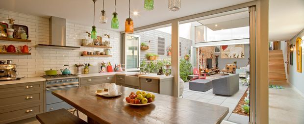 Bright, open-plan kitchen with a large timber island dining table, dark built-in cabinetry, white subway-tile splashback, open shelving, pendant lights, and extensive glazing opening toward a landscaped courtyard. A stairway and adjacent circulation space are visible on the right.