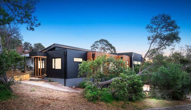 Single-storey contemporary dwelling set within a natural, lightly landscaped bush setting. The building has dark horizontal cladding, simple low-pitched roof forms, timber-toned wall sections, large rectangular windows, and a sheltered entry porch.