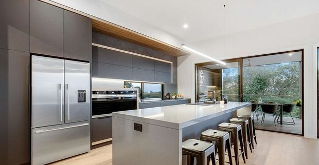 Contemporary open-plan kitchen with dark matte full-height cabinetry, integrated stainless-steel refrigerator and ovens, a large pale stone island with seating for five, and warm timber detailing. Floor-to-ceiling glazed doors open to an outdoor dining area and greenery beyond.