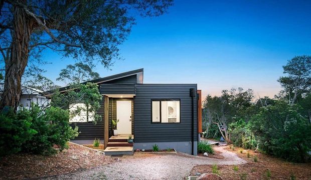 A compact single-storey modern home set among established trees and landscaped vegetation. The dark horizontal cladding, simple projecting roof forms, large windows and timber-toned entry create a contemporary cabin-like facade, with a gravel path and small planted garden in the foreground.