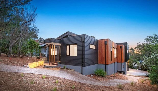 A modern single-storey house set on a sloping, wooded site. The compact building has dark horizontal cladding, contrasting warm timber-clad volumes, a low pitched roof, large rectangular windows, and a small covered entry porch. Gravel paths and retaining elements surround the structure.