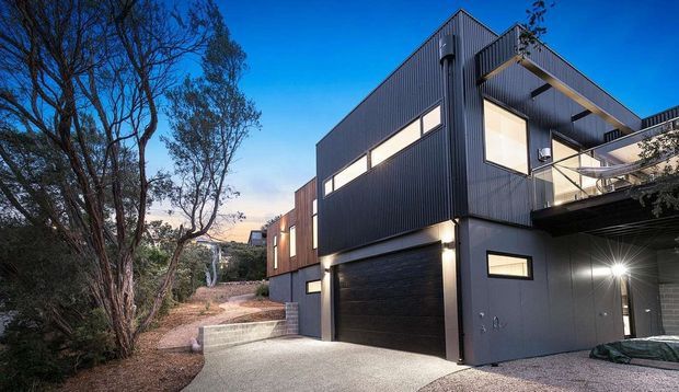 Exterior view of a contemporary multi-level home on a wooded, sloping site. The building has dark vertical metal cladding, large horizontal windows, projecting balconies, a flat roof, and an integrated double garage. A concrete driveway and retaining wall lead up through mature trees.