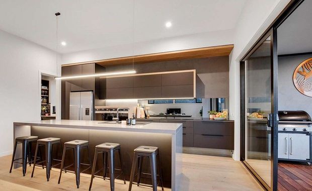 Contemporary open-plan kitchen with a large island and four metal bar stools, dark brown built-in cabinetry, a pale stone benchtop, integrated appliances, timber accents, and a white ceiling with recessed lighting. Full-height glazed doors open to an outdoor barbecue area, visible on the right.