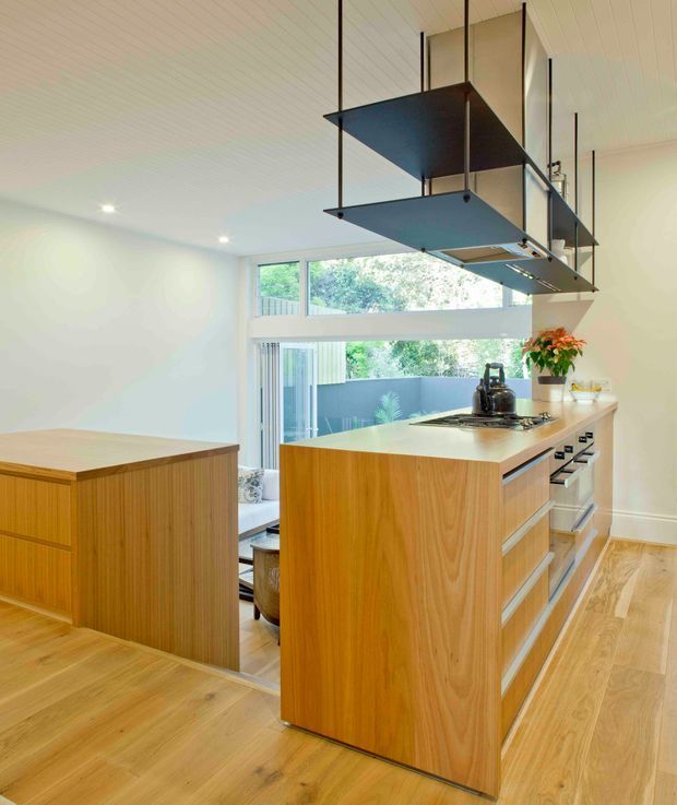 Bright contemporary kitchen with warm timber cabinetry and matching engineered timber flooring. A central island includes an integrated cooktop and built-in oven appliances, while a second timber peninsula or cabinet block sits in the foreground. White walls and a white-lined ceiling create a light backdrop, with large windows and glazed doors providing views to greenery and an outdoor courtyard. A distinctive suspended dark metal rangehood canopy hangs above the island.