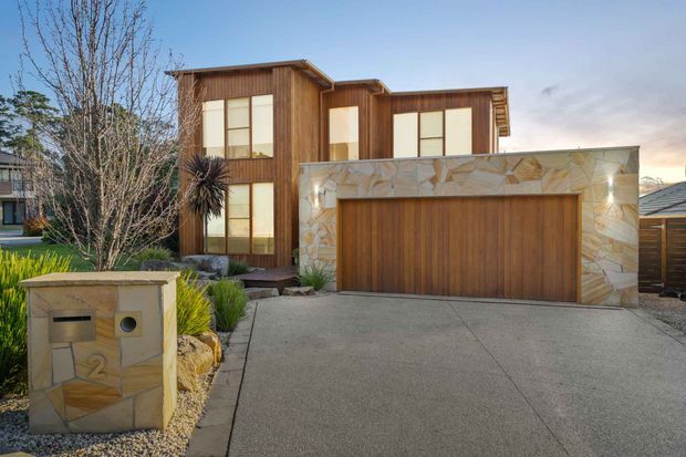 Exterior photograph of a contemporary two-storey residence with a warm timber-clad upper volume, large vertical windows, and a prominent double garage integrated into a pale irregular stone-faced structure. A broad concrete driveway, landscaped front garden, mailbox, and neighbouring homes are visible.