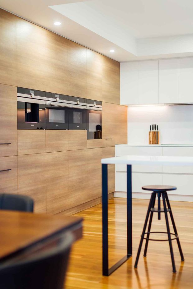 A contemporary kitchen featuring a full-height light timber joinery wall with integrated black appliances, white overhead cabinetry, and a large white island or breakfast bar supported by dark metal legs. Timber flooring, recessed ceiling lighting, and clean handleless cabinetry create a bright, refined minimalist interior. A black bar stool and part of a dining table are visible in the foreground.