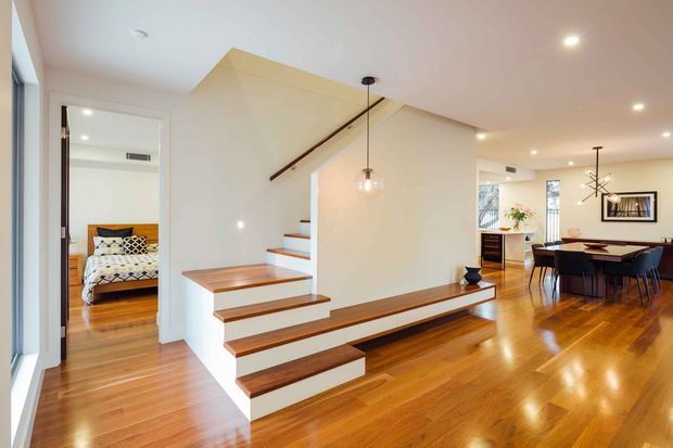 Bright open-plan interior featuring a timber staircase with white risers, integrated timber bench seating beneath the stairs, polished timber flooring, and a dining area with a dark table and chairs. A bedroom is visible through the doorway, while contemporary pendant and ceiling lighting illuminate the space.