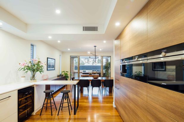 Bright contemporary open-plan kitchen and dining area with warm timber flooring and a full-height timber veneer appliance wall. The kitchen includes a white island with an integrated dark benchtop, stools, built-in black ovens, and a wine refrigerator. Beyond the island is a dining table with black chairs, large glazed doors or windows, and views to an outdoor area. Recessed ceiling lighting and clean-lined joinery create a polished, spacious interior.