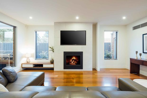 Bright contemporary living room with a large dark leather sectional sofa in the foreground, polished timber flooring, a central tiled fireplace with built-in firebox and wall-mounted television, expansive windows, and a dark timber console along the right wall. The room has a clean, open layout with recessed ceiling lights and neutral finishes.