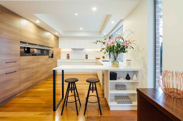 Bright contemporary kitchen with extensive warm timber cabinetry and flooring, a white island with integrated open shelving, two black bar stools, white benchtops, and a bank of built-in appliances. Recessed ceiling lighting and a tall glazed opening create an airy, refined space. A timber console with decorative objects is visible at right.