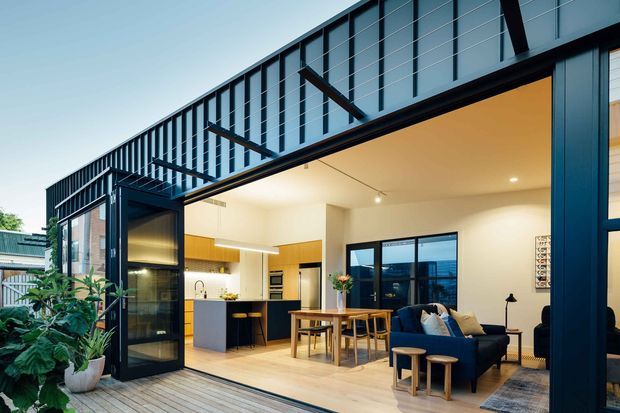 A contemporary open-plan ground-floor living space photographed from a timber deck, with a compact kitchen, dining table, and lounge arranged beneath a large covered opening. The home features extensive black-framed glazing and doors, a dark metal upper-level screen or balustrade, warm timber cabinetry and flooring, white walls, and indoor tropical plants. The partially visible exterior deck and neighbouring structures frame the scene.