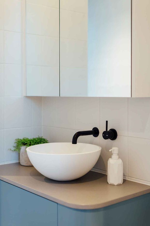 Close-up view of a contemporary bathroom vanity featuring a white vessel basin, matte black wall-mounted tapware, pale tiled walls, and blue-grey fixed cabinetry with a light neutral benchtop. A mirrored cabinet and small plant are also visible.