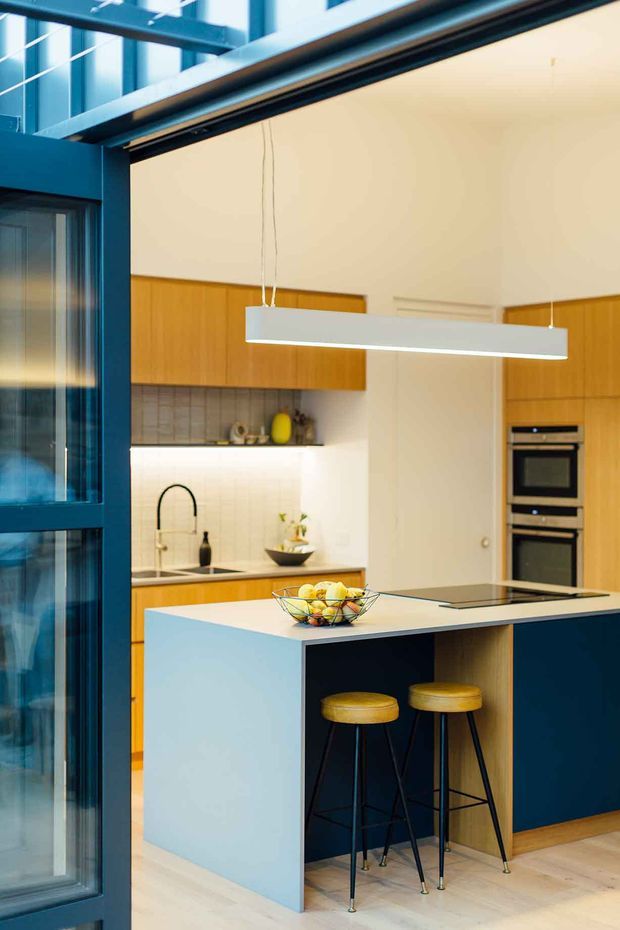 A bright contemporary kitchen viewed through a dark blue framed glazed opening. The room features a large pale blue island with an integrated dark blue seating recess, two timber-seated bar stools, light timber cabinetry, a pale tiled backsplash, integrated black ovens, and a white linear pendant light. A sink and cooktop are visible, with warm timber flooring and a high ceiling with exposed dark framing.