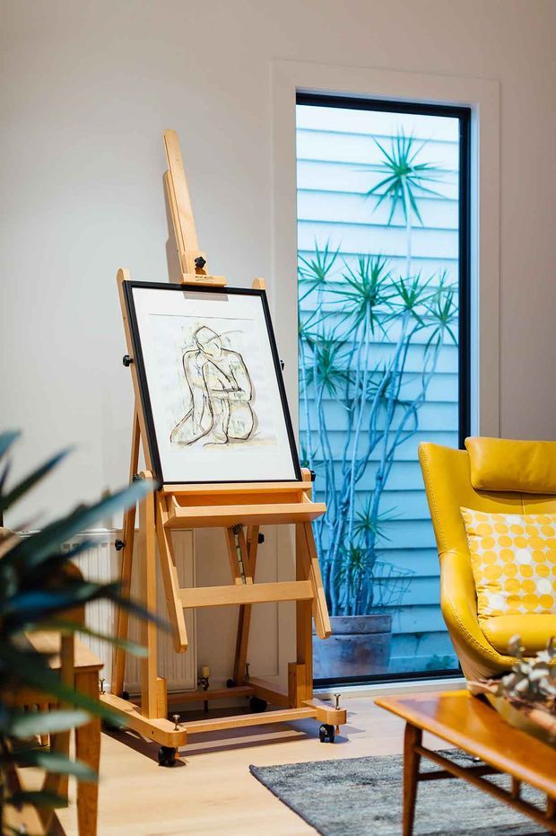 A warmly lit interior corner featuring a wooden artist's easel with framed artwork, positioned beside a tall black-framed window. A vibrant mustard-yellow armchair with a patterned cushion sits to the right, with a timber coffee table and rug partially visible. Plants and an exterior rendered wall are seen through the glazing, while the room has pale walls and light timber flooring.