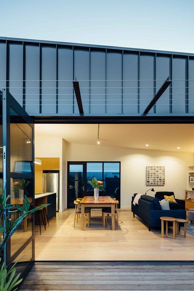 A contemporary open-plan living and dining space viewed from a timber deck through fully opened glazed doors. The room features pale timber flooring, a timber dining table and chairs, a dark blue sectional sofa, a compact kitchen with timber cabinetry, and warm recessed and pendant lighting. The upper facade has dark vertical metal cladding with exposed diagonal structural braces, while large glazing connects the interior to the deck and exterior view.